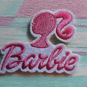 May include: A pink embroidered patch featuring the silhouette of a woman's head with a ponytail and the name "Barbie" in cursive. The patch is on a light grey background.
