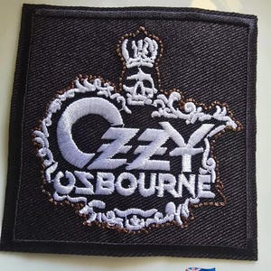 May include: Black square patch with the name "OZZY OSBOURNE" embroidered in white and brown. The design includes a skull with a crown and ornate scrollwork. The patch is likely for a jacket or bag.