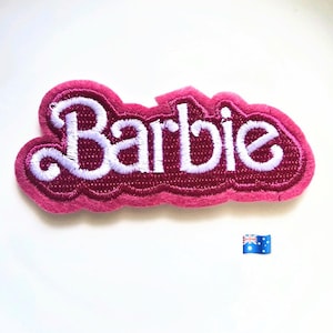 Puede incluir: Un parche de tela bordado que presenta la palabra Barbie en letras cursivas blancas. El texto está delineado con un borde grueso de color rosa oscuro que sigue la forma de las letras, creando una apariencia audaz y en relieve.