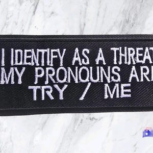 May include: A rectangular black embroidered patch featuring white stitched text that reads I IDENTIFY AS A THREAT MY PRONOUNS ARE TRY / ME. The patch has a thick black border and a textured fabric surface with clear, bold lettering throughout the design.