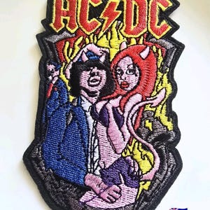May include: An embroidered fabric patch featuring the AC/DC logo in red and yellow at the top. Below the text, the design depicts a figure in a blue suit and a red-haired devil character with horns and a tail, set against a yellow flame background.
