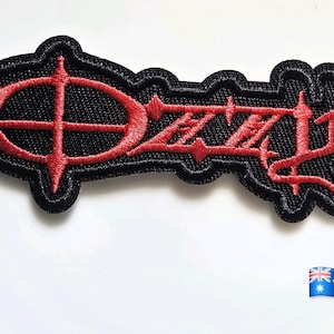 May include: An embroidered patch featuring a black background with red thread forming a stylized oval symbol on the left and the text OZZY in a sharp, angular font on the right. The patch has a thick black border and a textured fabric surface.