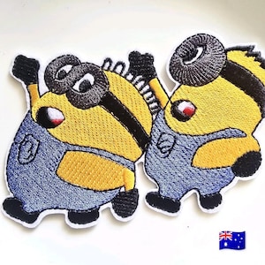Puede incluir: Dos parches bordados que muestran personajes de dibujos animados amarillos con gafas negras, petos azules y guantes negros. Los personajes aparecen en una pose dinámica con los brazos levantados, realizados con hilo amarillo, azul, negro y blanco.
