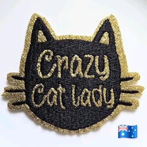 Op de afbeelding: Een katvormige patch met een zwarte, getextureerde achtergrond en een gouden glitterrand. De tekst Crazy Cat Lady staat in gouden glitterletters in het midden. De patch heeft puntige oren en drie snorharen aan elke kant van het gezicht.