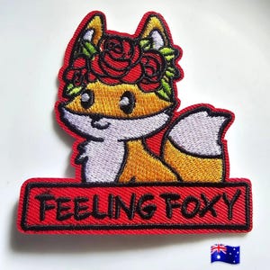 May include: Embroidered patch of a cartoon fox wearing a floral crown. The fox is orange, white, and yellow, with a red border. The patch includes the text "FEELING FOXY" in black letters on a red background.