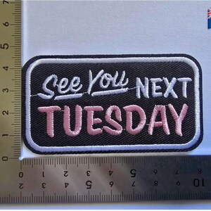 Puede incluir: Parche bordado con el texto "See You NEXT TUESDAY" en letras blancas y rosas sobre fondo negro. El parche tiene un borde blanco y una forma rectangular. Una regla está en la imagen.