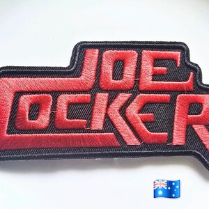 以下が含まれることがあります： 黒い背景に赤い刺繍の文字で「JOE COCKER」と書かれた長方形の刺繍ワッペン。文字は太いブロック体でデザインされており、刺繍されたデザインの形状全体が厚い黒い縁取りで囲まれています。