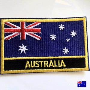 May include: A rectangular embroidered patch featuring the Australian flag with a blue field, the Union Jack in the upper left corner, and white stars. A black horizontal band at the bottom displays the word AUSTRALIA in gold embroidery.