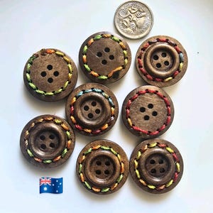 May include: Nine circular wooden buttons with four holes, each with colourful embroidered edging. A silver coin and a small Australian flag are also present. The buttons are a medium brown colour and the embroidery is in various colours.