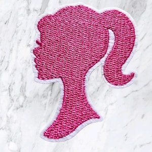 Peut inclure: Un écusson brodé rose vif représentant la silhouette d'une tête de profil avec une queue de cheval haute. Le motif est souligné par une épaisse bordure blanche, mettant en valeur un point texturé sur toute la forme rose éclatante.