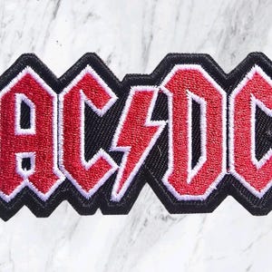 May include: An embroidered patch featuring the text AC/DC in a bold, block-style font. The letters are red with a white outline, set against a solid black background. The central lightning bolt symbol separates the letters AC and DC in red.