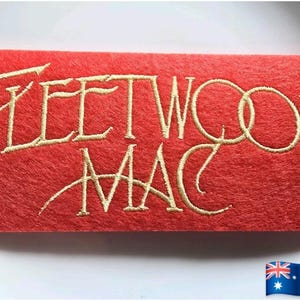 Op de afbeelding: Een rechthoekig, rood vilt item met de geborduurde tekst "FLEETWOOD MAC" in een goudkleurig script. Het item is waarschijnlijk een patch of een decoratief stuk, mogelijk voor een tas of kleding. De Australische vlag staat rechtsonder.