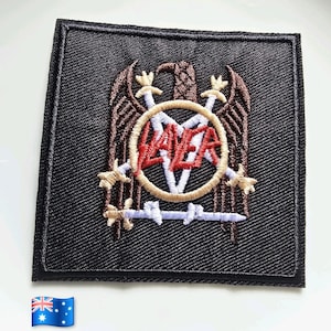 May include: A square black embroidered patch featuring a brown eagle with spread wings, two crossed swords, and a central circle containing the red text Slayer. The design includes a light blue barbed wire element across the bottom section.
