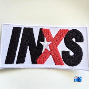 May include: A rectangular embroidered patch featuring the text INXS in a bold, sans-serif font. The letters I, N, and S are stitched in black thread, while the letter X is stitched in red thread with a small white star centred on it.