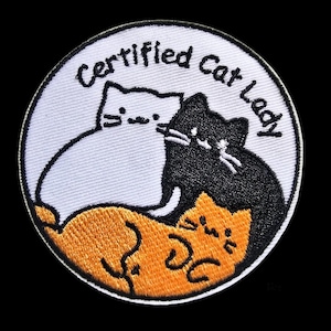 May include: A circular embroidered patch with a white background featuring three cats in white, black, and orange. The text "Certified Cat Lady" is stitched in black along the top curve of the patch, which has a thick black border.