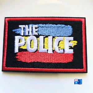 Puede incluir: Un parche rectangular bordado con fondo negro y borde rojo. El texto THE POLICE está bordado en blanco, superpuesto sobre franjas horizontales de hilo azul claro, amarillo y rojo que imitan pinceladas en el centro.