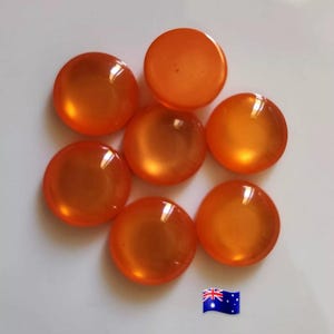 May include: Seven translucent, orange, round cabochons are arranged on a white surface. The cabochons have a glossy finish and reflect light. The Australian flag is in the bottom right corner. These cabochons are suitable for jewellery making or craft projects.