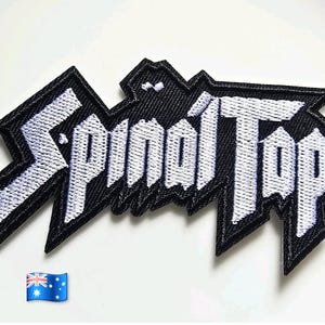 May include: A black and white embroidered patch featuring the text Spinal Tap in a jagged, stylised font. The design has a thick black border and white lettering with sharp, angular edges, creating a bold, high-contrast appearance for this fabric accessory.