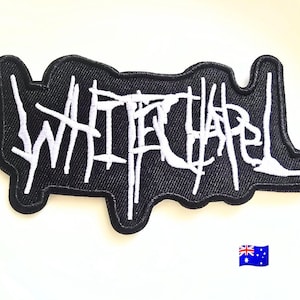 Puede incluir: Un parche bordado negro con la palabra Whitechapel en letras blancas estilizadas y dentadas. El parche tiene un borde negro grueso y una forma orgánica irregular que sigue los contornos del texto. Se puede ver una pequeña bandera de Australia.