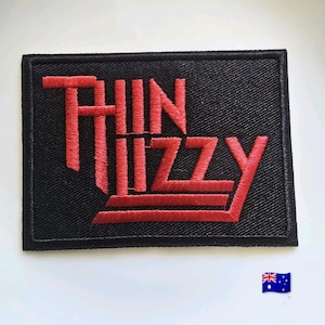 Puede incluir: Un parche bordado rectangular con fondo negro y el texto rojo Thin Lizzy cosido en una fuente estilizada. El parche tiene un borde negro cosido y una forma rectangular, mostrando el logotipo de la banda en hilo de color rojo intenso.