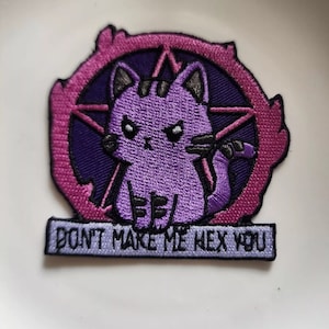 May include: Embroidered patch with a purple cat inside a pentagram, set against a pink and purple border. The cat has a stern expression. The text "DON'T MAKE ME HEX YOU" is displayed below the cat.
