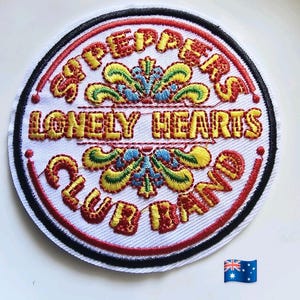 May include: Circular embroidered patch featuring the text "Sgt. Peppers Lonely Hearts Club Band". The design uses yellow and red thread on a white background, with a black and red border. Decorative floral elements are also present.