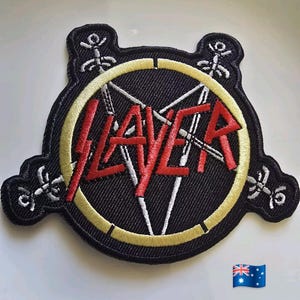 Könnte beinhalten: Gestickter schwarzer Aufnäher mit dem Wort "SLAYER" in Rot, einem gelben Rand und einem weißen Pentagramm-Design. Der Aufnäher hat eine einzigartige Form mit dekorativen weißen Akzenten an den Rändern.