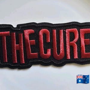 May include: Embroidered patch with the text "THE CURE" in bold, red letters. The letters are stitched onto a black denim-like background. The patch has a black border and is likely designed to be ironed or sewn onto clothing or accessories.