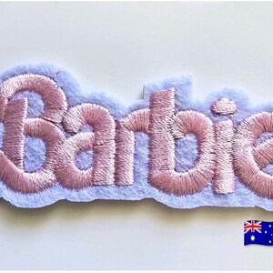 May include: A fabric patch featuring the word Barbie embroidered in a light pink thread with a satin finish. The lettering is outlined and set against a light lavender felt backing that follows the contour of the script text. A small Australian flag icon is visible in the bottom right corner.