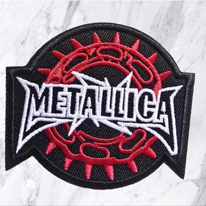 Puede incluir: Un parche bordado circular con fondo negro y un diseño de corona de espinas roja. El logotipo blanco de Metallica está bordado en el centro, con las letras M y A extendiéndose hacia afuera en puntas afiladas y angulares en los lados.