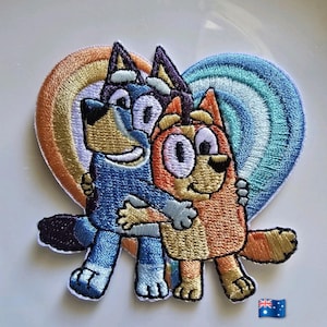 May include: An embroidered patch featuring two cartoon dogs, one blue and one orange, hugging in front of a heart-shaped background with rainbow-colored stripes. The blue dog is on the left and the orange dog is on the right, both smiling.