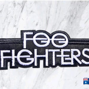 May include: A rectangular embroidered patch featuring the text "FOO FIGHTERS" in bold white capital letters against a black background. The design includes a stylised border and white stitching that outlines the letters, creating a high-contrast look.