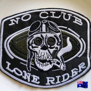 May include: A black embroidered patch featuring a white skull wearing aviator goggles and smoking a cigar. The text NO CLUB is arched above the skull, and LONE RIDER is arched below. The design is set against an olive green oval background.