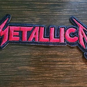 May include: Embroidered patch with the word "METALLICA" in red, with a lightning bolt design. The patch has a dark blue denim-like background and a black border. Suitable for application on clothing or accessories.