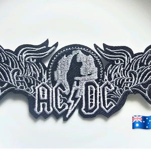 Puede incluir: Un parche bordado en blanco y negro que presenta el texto AC/DC con un rayo en el centro. El diseño incluye formas estilizadas similares a alas que se extienden desde un emblema circular central. Un pequeño icono de la bandera australiana es visible en la esquina.