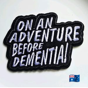 Peut inclure: Écusson brodé noir avec les mots "ON AN ADVENTURE BEFORE DEMENTIA!" en fil blanc. Le patch a une bordure noire et une forme stylisée. Le drapeau australien est dans le coin inférieur droit.