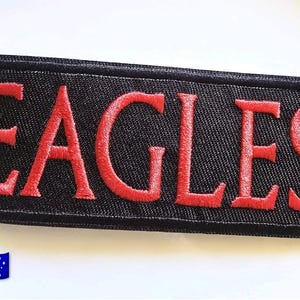 Può includere: Una toppa rettangolare ricamata con sfondo nero e un bordo nero spesso. La parola EAGLES è cucita in lettere maiuscole rosse e in grassetto al centro della toppa. Il carattere presenta uno stile classico con grazie.