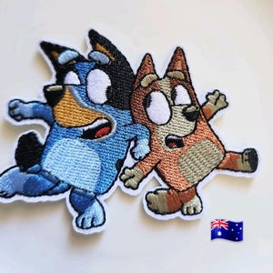 Puede incluir: Parches bordados con dos perros de dibujos animados, uno azul y otro marrón, en poses dinámicas. Los parches tienen bordes blancos y están diseñados para aplicarse en ropa o accesorios. Una bandera australiana es visible.