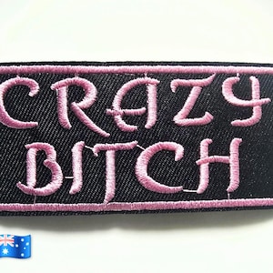 May include: A rectangular embroidered patch with a black background and a pink border. The text CRAZY BITCH is stitched in pink thread in the centre of the patch. The patch features a textured fabric surface and a clean, finished edge.