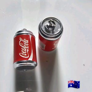 Puede incluir: Dos recipientes en miniatura con forma de lata de Coca-Cola. Las latas son rojas con el logotipo de Coca-Cola en escritura blanca. Las tapas y los fondos son plateados. Una lata está cerrada y la otra está abierta, mostrando la anilla. El fondo es blanco.