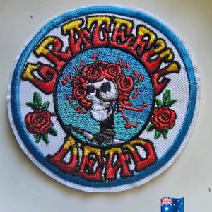 May include: Circular embroidered patch with the words "Grateful Dead" in a decorative font. The design features a skull with roses, set against a blue background, with a blue and white border. Red roses are also included.