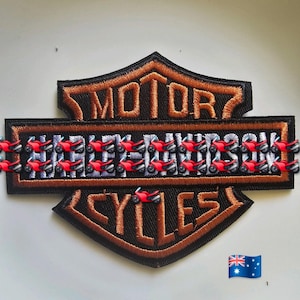 May include: Black and brown embroidered patch with the words "MOTOR HARLEY-DAVIDSON CYCLES". The patch has a shield shape and is decorated with small red motorcycle charms. An Australian flag is also present.