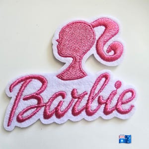May include: An embroidered patch featuring the pink Barbie logo in a cursive script font positioned below a pink silhouette of a head with a high ponytail. The design is stitched onto a white fabric backing with a scalloped edge finish.