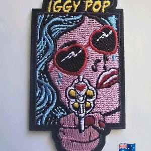 以下が含まれることがあります： イギー・ポップの様式化された肖像画が刺繍されたパッチ。デザインには、ピンク色の肌、青い髪、ハート型のサングラス、リボルバーが含まれています。「IGGY POP」の文字が上部にあります。パッチには黒い縁取りがあります。