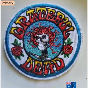 Könnte beinhalten: Runder, gestickter Aufnäher mit den Worten "Grateful Dead" in roter und gelber Schrift. Das Design zeigt einen Totenkopf mit roten Rosen, einen blauen Hintergrund, einen blauen Rand und weißen Stoff.
