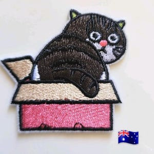 May include: Embroidered patch of a cartoon cat in a pink box. The cat is brown and white with large eyes and a pink nose. The box is pink with a beige top. The patch has a white border.