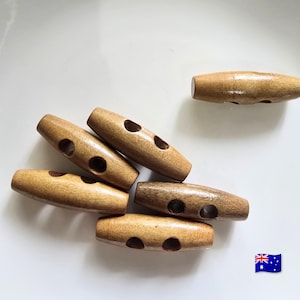 May include: A collection of six light brown wooden toggle buttons, each featuring a smooth, cylindrical shape with two holes drilled through the center for attachment. The buttons have a natural wood grain finish and are arranged in a cluster.
