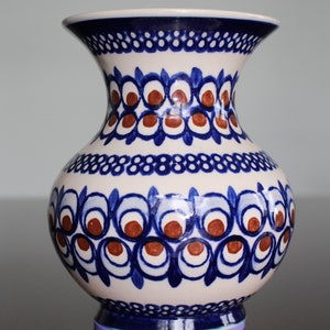 Könnte beinhalten: Eine weiße Keramikvase mit einem blauen und braunen Blumenmuster. Die Vase hat eine breite Öffnung und einen runden Körper. Sie hat einen violetten Sockel.