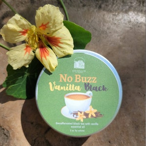 May include: A round tin of decaffeinated black tea with vanilla essential oil. The tin is green with white text that reads "No Buzz Vanilla Black". A small cup of tea and vanilla beans are pictured on the tin.