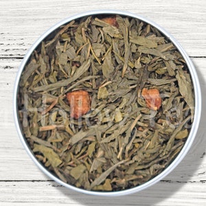 May include: A round metal tin filled with loose leaf green tea and dried fruit pieces. The tea is a mix of green leaves and small pieces of dried fruit.